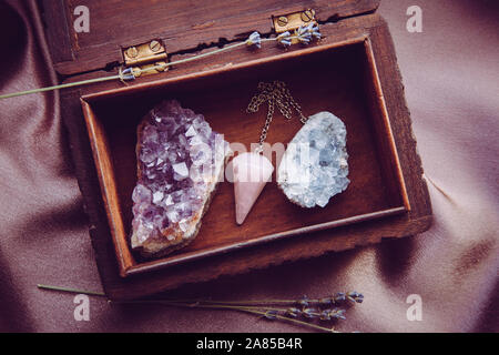 Witch tools inside beautiful old wood box. Rose quartz pendulum, natural amethyst and celestite crystal clusters. Dry lavender flowers on dark purple Stock Photo
