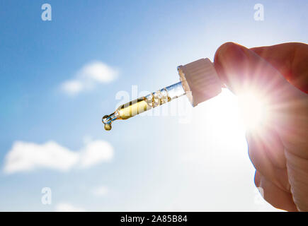 Hand holding dropper pipette with nice golden liquid D-vitamin against sun and blue sky on sunny day. Vitamin D keeps you healthy while lack of sun in Stock Photo
