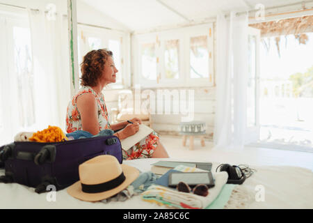 Serene woman writing in journal next to suitcase in beach hut bedroom Stock Photo