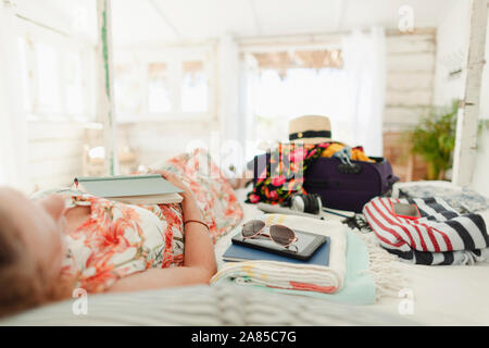 Woman with book relaxing on bed next to suitcase in sunny beach hut bedroom Stock Photo
