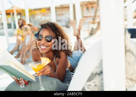 Portrait happy, carefree young woman reading book and drinking cocktail on beach Stock Photo