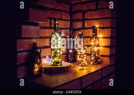 Lot of different vintage style beer bottles and green wine bottles decorated with wire string micro led lights on shelf, red brick wall background. Il Stock Photo