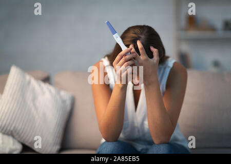 Desperate woman holding pregnancy test sitting on sofa at home Stock Photo
