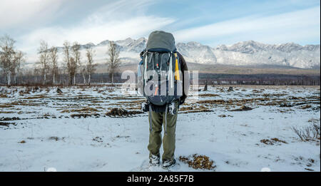 A man hiker with a large backpack stands with his back looking at the snowy mountains ahead. Concept the beginning of the path to achieving the goal Stock Photo