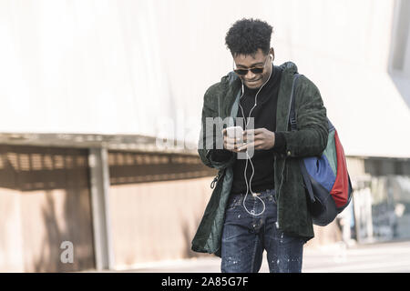 black young man with earphones walking by the city and texting with a mobile phone, technology and lifestyle concept using internet electronic device Stock Photo