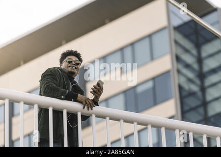 portrait of a smiling young black man with sunglasses listening to music with earphones and mobile phone while leaning on a handrail outdoors in the c Stock Photo