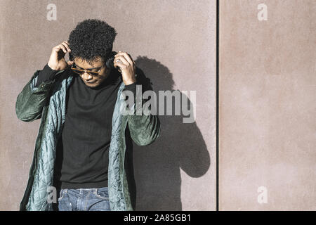 cool african black man with casual modern clothes and headphones listening music leaning against wall, technology and lifestyle concept Stock Photo