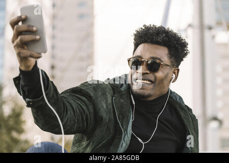 handsome young african black man with sunglasses making a video call with smartphone and earphones while sitting in the city, lifestyle and technology Stock Photo
