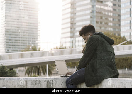 modern young man with earphones working on laptop sitting outdoors in the city, lifestyle and technology concept using internet electronic device Stock Photo