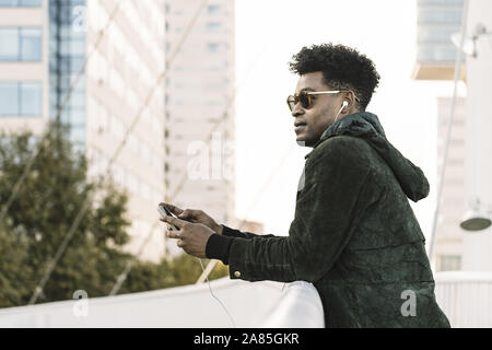 cool young african black man with sunglasses listening music on smartphone with earphones while leaning on a handrail outdoors in the city, lifestyle Stock Photo
