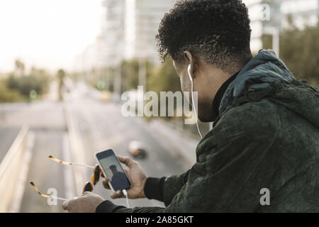 view from behind of a young african black man with sunglasses making a video call with smartphone and earphones, city lifestyle and technology concept Stock Photo