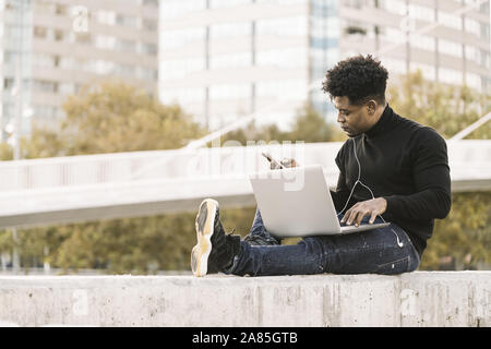 handsome black student with white earphones working with computer and smart phone sitting outdoors in the city at sunset, lifestyle and technology con Stock Photo