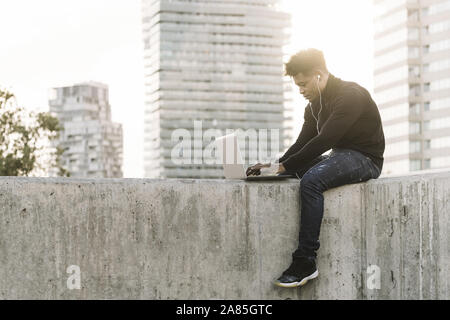 handsome black man with white earphones working typing in a computer sitting outdoors in the city at sunset, lifestyle and technology concept using in Stock Photo