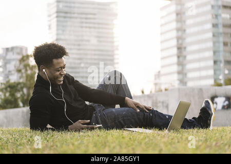 smiling young black man laughing with white earphones looking to the laptop lying on the lawn in the city at sunset, lifestyle and technology concept Stock Photo