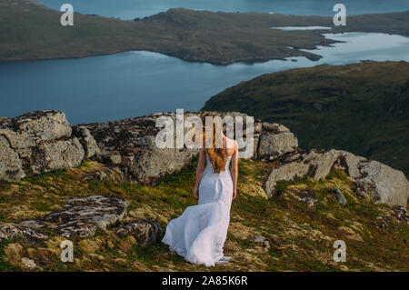 Back of girl in wedding dress in mountains looking to fjord, hiking in hills Stock Photo