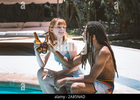 Stock photo of a multiracial couple sitting on the edge of the pool toasting with beer and smiling with the garden in the background. Summertime Stock Photo