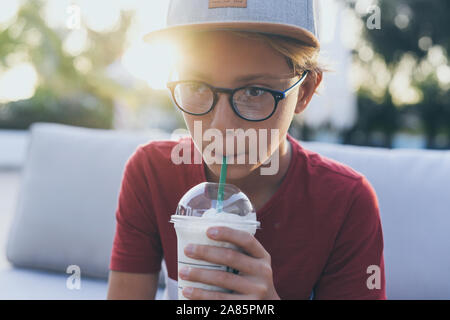 Trendy young boy drinking a smoothie sitting outdoor at the bar. Teen with hat drinks a cold beverage outside in the garden. Portrait of pensive teena Stock Photo