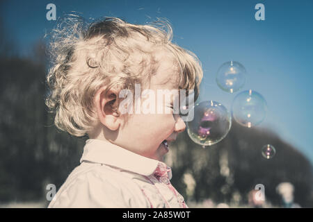 Beautiful little haired hair girl, has happy fun smiling face, pretty eyes, short hair, playing and catching soap bubbles in summer nature, dressed Stock Photo