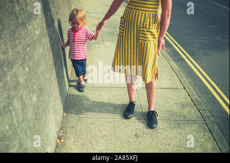 A young mother and her toddler are walking on the street by a wall in summer Stock Photo