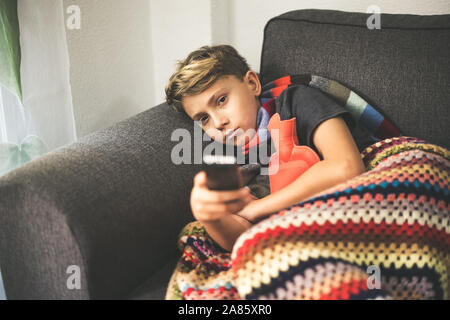 Sick boy watching tv with woolen blanket and hot water bottle. Sad teen with the flu rests at home in a cold winter day. Child with seasonal infection Stock Photo