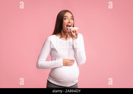 Pregnant Woman Eating Donut On Pink Background, Studio Shot Stock Photo