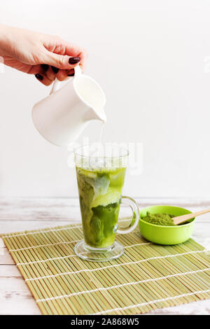 Iced matcha latte drink in tumbler glass with coconut milk pouring from pitcher by hand, copy space. Summer refreshing vegan beverage cold drink. Stock Photo