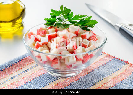 Sliced surimi crab sticks in a glass bowl on a striped table mat near olive oil glass jar and chef knife. Seafood and ingredient for salads. Stock Photo