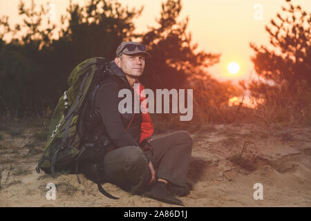 Tired mature hiker with big backpack sit down on sand against sunset light in forest. Stock Photo