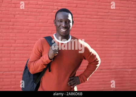 Portrait Of African Young Man Looking At Camera And Smiling, Carrying Backpack For School. Copy Space On Red Wall In Background. Stock Photo