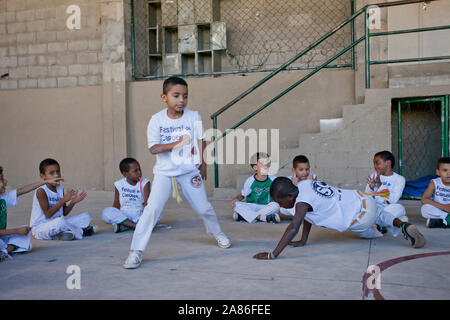 Kids at a Brazilian capoeira class with a teacher playing a berimbau with their peers in a circle in the background at an outdoor sports venue Stock Photo