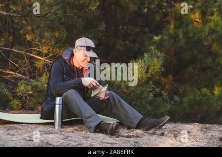 Elderly hiker eating a sandwich and drinking a hot drink sitting on a tourist rug in forest. Rays of sunset illuminates a man. Stock Photo