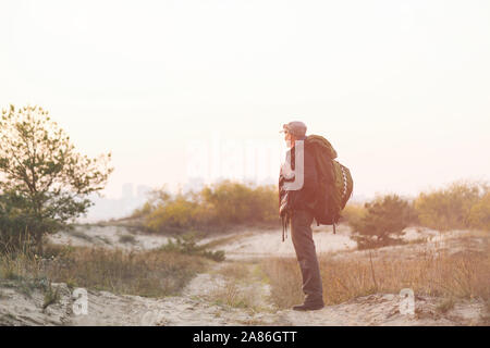 Tourist with big green backpack looking far. A man walks along a sandy road. City in fog haze in background. Stock Photo
