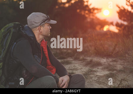 Man looks at beautiful sunset. Tired mature hiker with big backpack sit down on sand against sunset light on sand at nature. Stock Photo