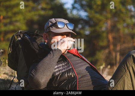 Tired tourist is resting in forest. Traveler man at halt drinks from a thermos. Stock Photo