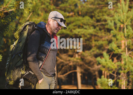 An aged man with backpack in tourist clothes, sunglasses and a cap. Forest in the background. Stock Photo