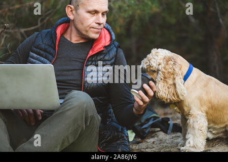 Older tourist using computer outdoors during travel with cocker spaniel at forest. Smiling man feeds a dog. Stock Photo