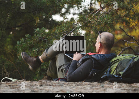 Elderly stylish man tourist use laptop in forest. Back view, male hiker lying on back leaning on backpack, with laptop computer, typing, blogging, bro Stock Photo