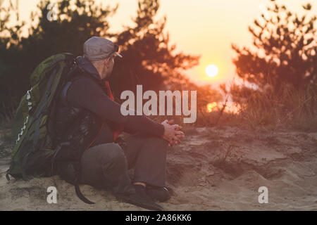 Man looks at beautiful sunset. Tired mature hiker with big backpack sit down against sunset light on sand at nature Stock Photo
