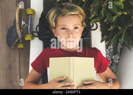 Thoughtful teen lying on a bench looking the sky with a book open on the chest. Young boy dreaming about fantastic stories after read a book. Kid Stock Photo