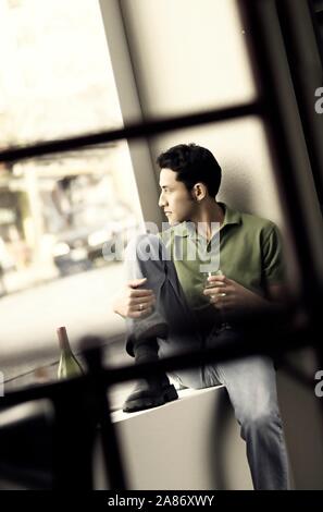 Asian man sitting alone in a large window with a glass of wine Stock Photo