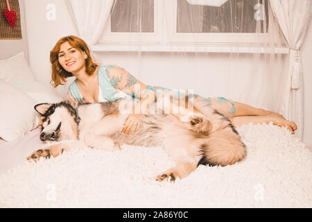 charming plus size girl with red hair in a nightgown posing with her large dog, a Malamute best friend in white bed in the bedroom. Stock Photo