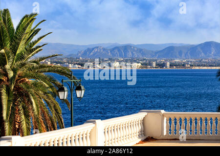 Beautiful seascape on the promenade, a street lamp under a palm tree looks at the sea, city and mountains in blur. Stock Photo