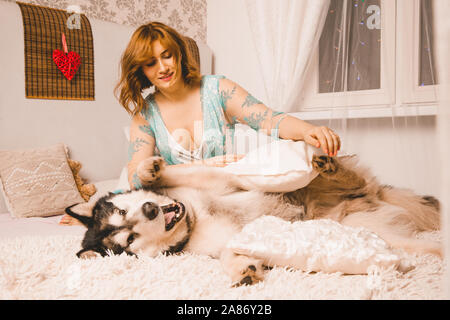 charming plus size girl with red hair in a nightgown posing with her large dog, a Malamute best friend in white bed in the bedroom. Stock Photo