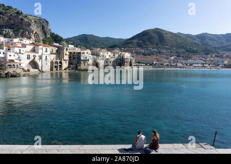 A couple sitting on the pier opposite the old town of Cefalú, Sicily, Italy, with the rocky crag of La Rocca looming over the town. Stock Photo