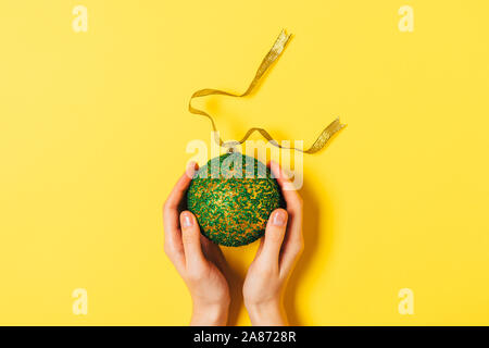 Female hands holding big Christmas ball on yellow background, top view. Stock Photo