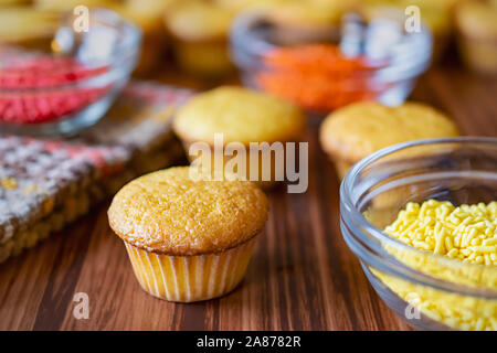 Three mini cupcakes with glass bowls of red, orange and yellow sprinkles on a wood surface. Stock Photo
