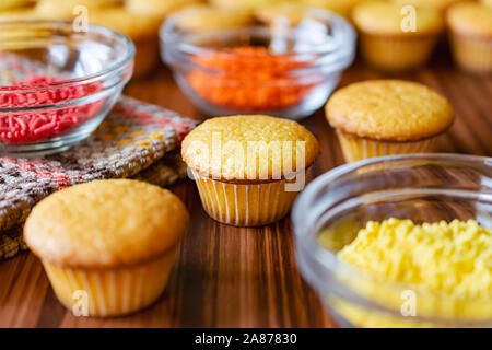 Three mini cupcakes with glass bowls of red, orange and yellow sprinkles on a wood surface. Stock Photo