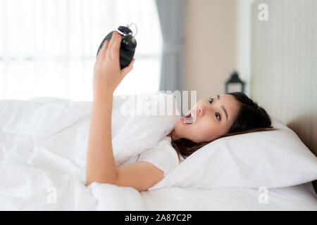 Beautyful Asian Woman lying on white bed and holding black alarm clock with shock to learn that late in morning at bedroom at home. Stock Photo