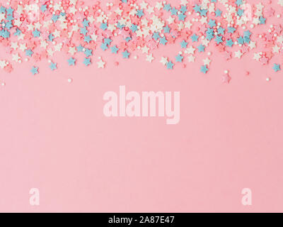 Festive border frame of colorful pastel sprinkles on pink background, copy space top. Sugar sprinkle dots and stars, decoration for cake and bakery. Top view or flat lay Stock Photo