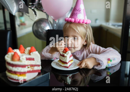 Indoor shot of pretty joyful little girl with blonde hair blowing out the candle, celebrate 6 years old birthday, wear fashionable dress, have excited Stock Photo